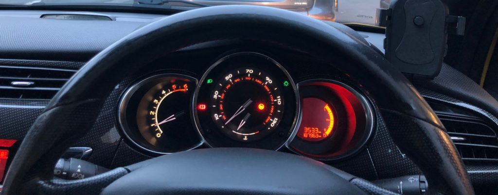 Front view of a car dashboard with three circle dials which are lit up. and the top half of the driving wheel.