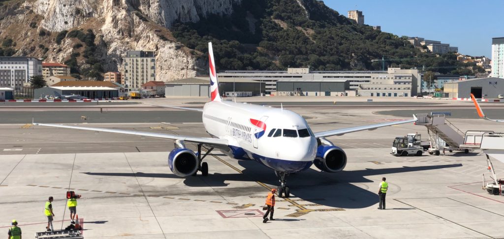 A British Airways A320 parked on the stand at Gibraltar Airport. There are some ground crew (wearing yellow and orange high-vis jackets) in the foreground and the Rock of Gibraltar is in the background.