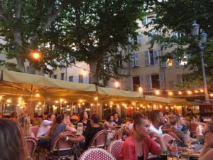 Pretty outside terrace in a French square, filled with parasols and fairy lights and lots of people eating and drinking at restuarant tables.