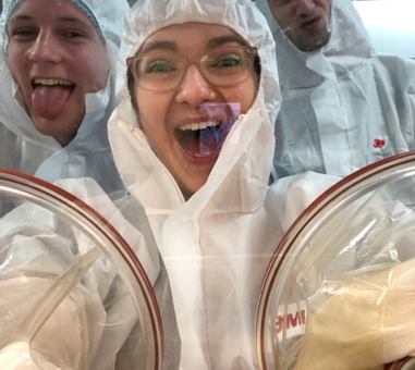 Three students wearing full-body aseptic suits taking a picture of themselves with the camera inside a sterile isolater while they're outside.