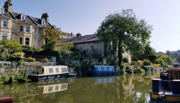 A photo taken of the Kennet and Avon canal. Georgian buildings overlook the narrowboats lining the banks full of lush vegetation on a bright sunny day.