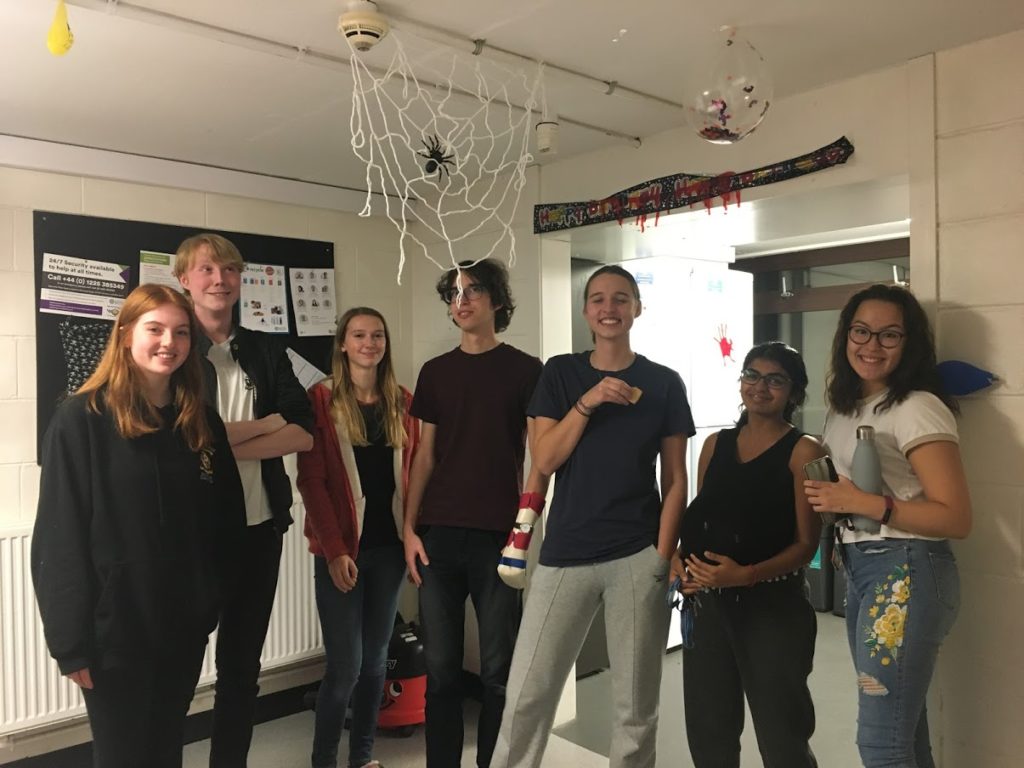 Seven people standing next to each other in a kitchen with white walls and ceiling, it is Halloween so there is a fake spiderweb with a fake spider on it hanging from the ceiling, there is an old Happy Birthday banner on the wall with fake blood on it. Everyone is smiling and posing, everyone is very cute.