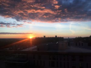 Sunset over buildings, visible from Norwood House. The clouds are lit by the orange sun rays and shadowed with purple. The little bit of visible sky is baby blue and yellow.