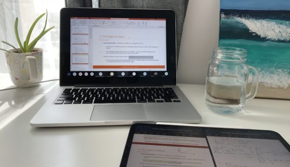 A picture of a desk set-up for studying. There's a laptop open with a tablet in front of it. There's a potted aloe vera plant to the left of the laptop and a glass of water to its right.