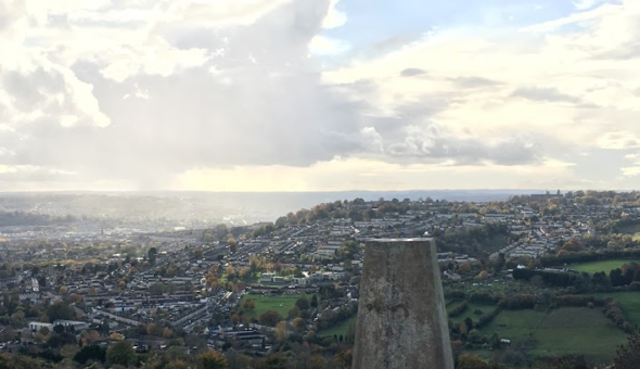 A grass covered hill on a sunny day, white fluffy clouds and a blue sky. In the middle of the picture, there's a small stone monument that looks like a cut pyramid. In the background, a city is visible (Bath) and the sun rays are falling right on it.