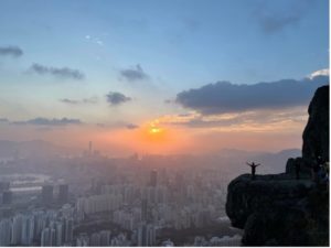 Student stood on the ledge of a mountain with arms outstretched. The city of Hong Kong in the background with a stunning sunset