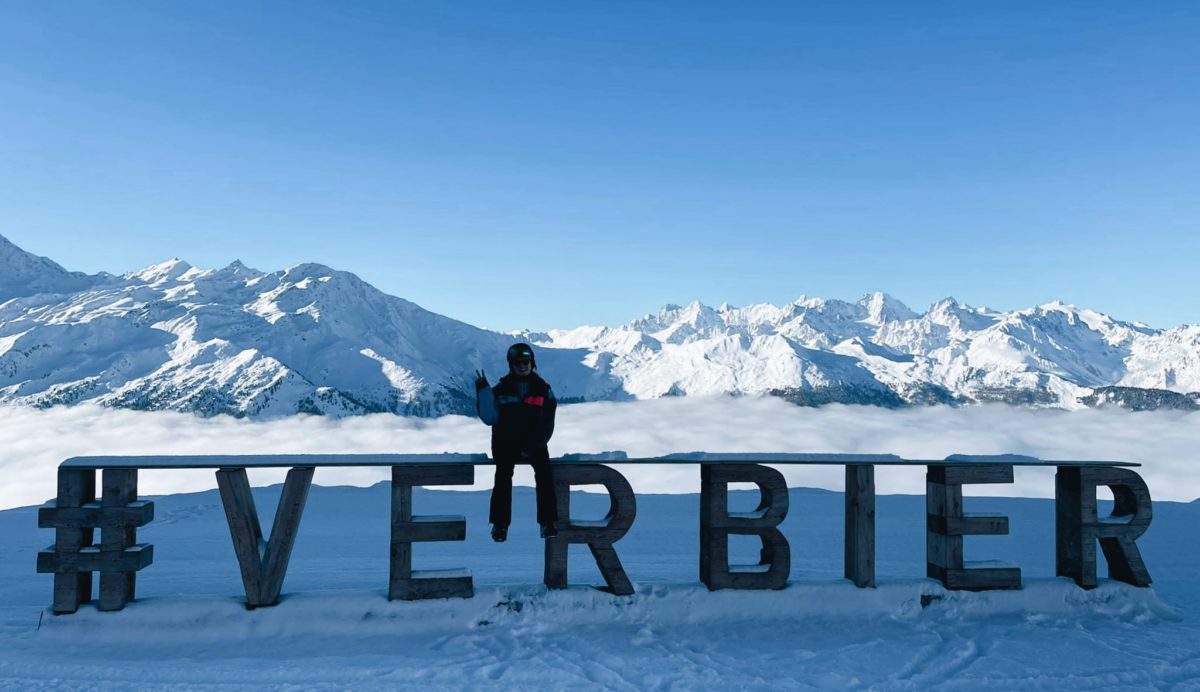 Me sat on a Verbier sign with snow covered mountains in the background in Verbier, Switzerland.