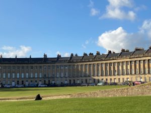 A view of the Royal Crescent with it's bath stone walls in a crescent shape, blue sky with a few clouds and green grass in the foreground