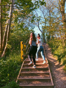 Two female student walking up some wooden stairs outside in some woods and looking over their shoulders.