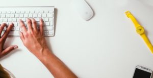 A pair of hands resting on a white keyboard with a white mouse top and centre, yellow watch middle,right and black notebook bottom, right.