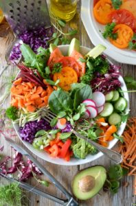 A very large bowl of colourful, prepared vegetables. There is half an avocado and kitchen utensils, used in the food preparation, outside the bowl and some sliced tomatoes in a separate bowl in the top right-hand corner. 