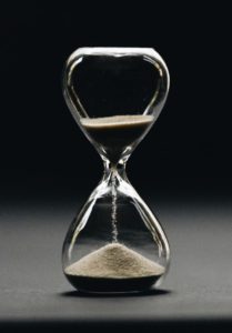 Photo of a glass sand timer with sand in both sections. The background graduates in colour from black to light grey 