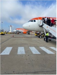 The front nose of an aeroplane on a runway with passengers boarding using the external steps. There is another orange and white aeroplane in the background. There is a zebra crossing in the foreground.