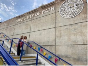 Me and my mum stood half way up some stairs with blue railings and colourful bunting on the railings. There is a grey wall behind us with the University of Bath and logo on the wall