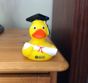 A bright yellow rubber duck with an orange beak, black mortar board, holding a white scroll with red ribbon.