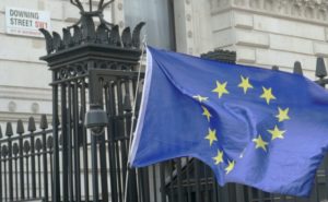 A circle yellow stars and blue background European Union flag blowing in the wind against the black metal railing and the Downing Street street sign