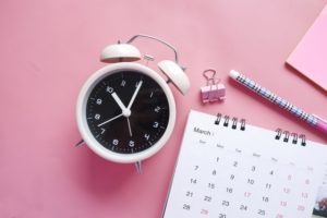 A white alarm clock, bulldog clip, pencil and calendar against a pink background