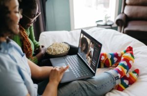 Two females sat on a bed in front of a laptop with a bowl of popcorn