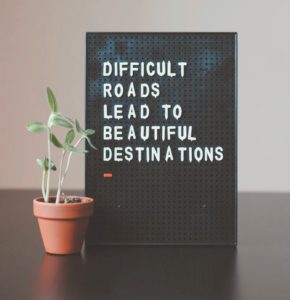 A black peg board which says Difficult roads lead to beautiful destinations in white test. With a brown pot plant with a green shoot next to it.