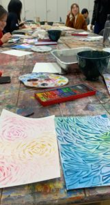 A table with artworks, paints and other art tools, with young people laughing and talking.