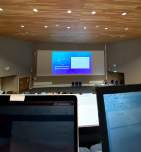 A view of a big screen in a lecture theatre with students working at their laptops.