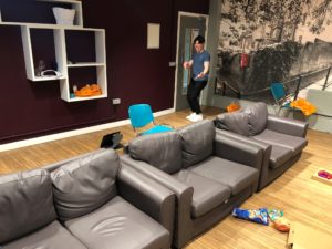 The lounge with a row of grey sofas on a wooden floor and white open shelving on the dark brown walls. There is a student stood in the doorway.