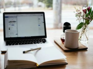 A laptop in the corner, with a notebook open in front and a pen placed on top. To the right, there is a white mug on a coaster and a plant on the edge of the shot. 