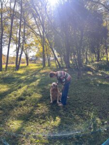 Woman (Nell) stood next to a medium sized dog, in a forest