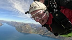 Close up of Grace's face, wearing a helmet and goggles, high above a large body of water 