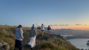 View of the back of five people (Grace and friends) as they walk across the top of a rocky hill, with the view of water and more hills in the distance
