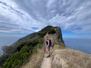Grace standing on a dirt path on a grassy hill, with the ocean in the background