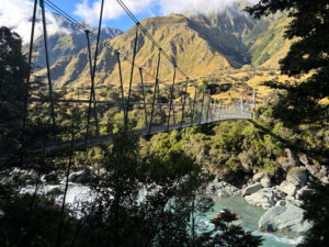 Large rope bridge across a river, with trees in the foreground and mountains in the background.
