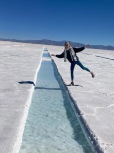 A person (Nell) stood on one leg next to a channel of clear water that runs through a salt flats, called the 'Las Salines Grandes’